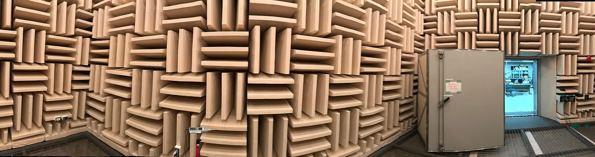Panorama of an acoustic anechoic chamber, its walls covered in sound-absorbing foam wedges, with a sound level meter on a stand and the door open to a lab