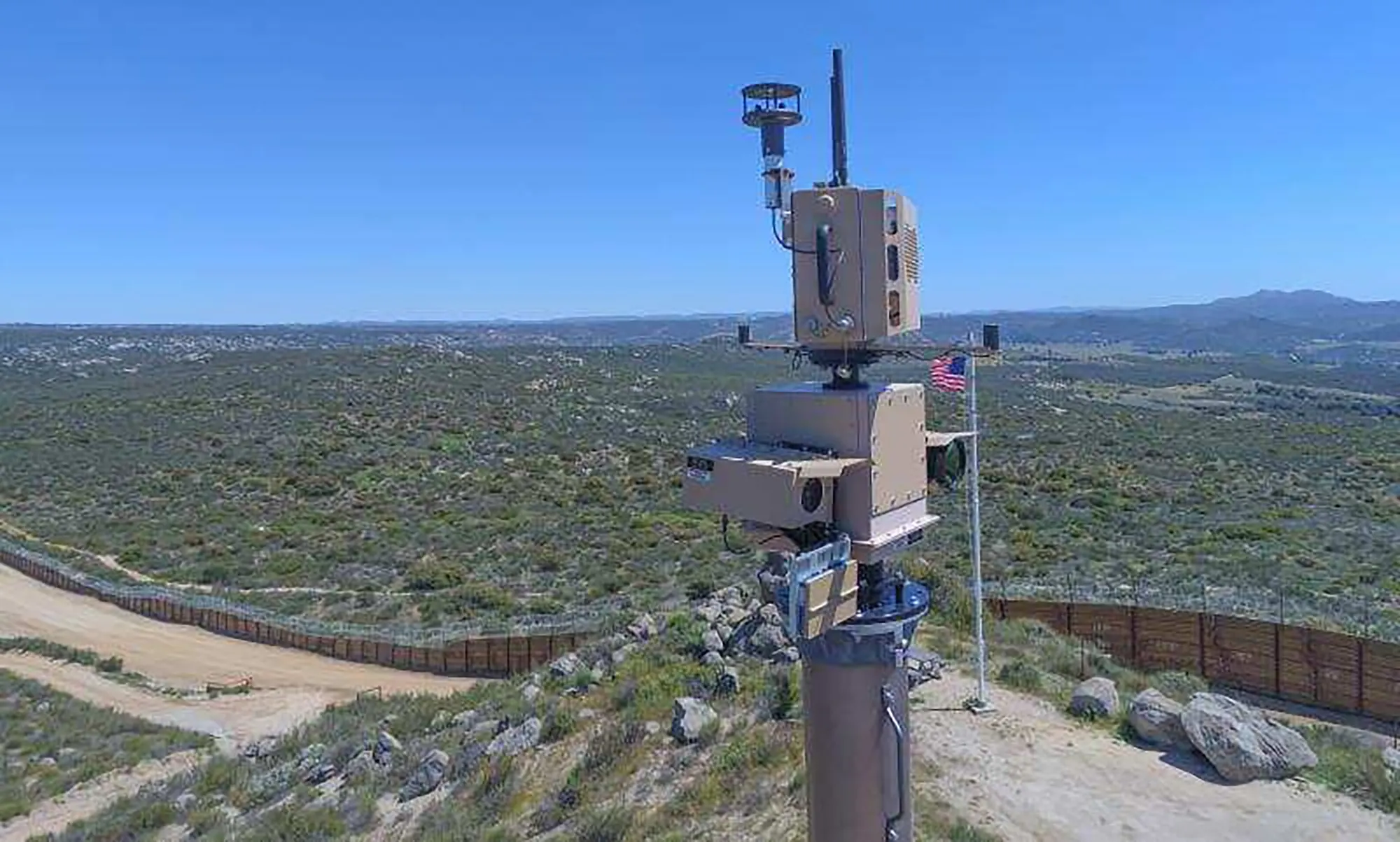 A CBP autonomous surveillance tower with a radar and camera head on a mast, overlooking scrubland and the border fence under a blue sky