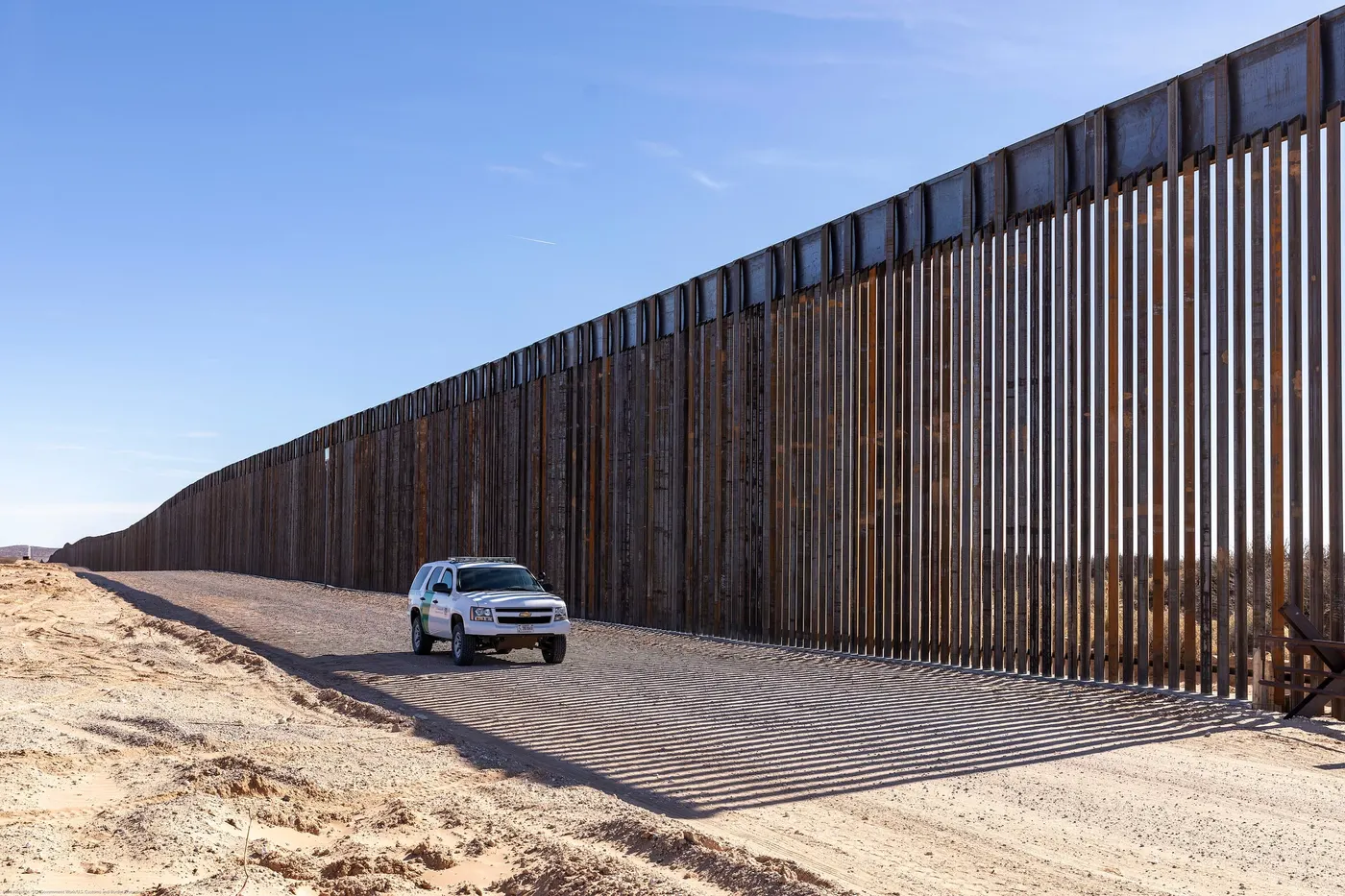 A tall steel bollard border wall running into the distance across sandy desert at Santa Teresa, New Mexico, with a Border Patrol vehicle on the service road