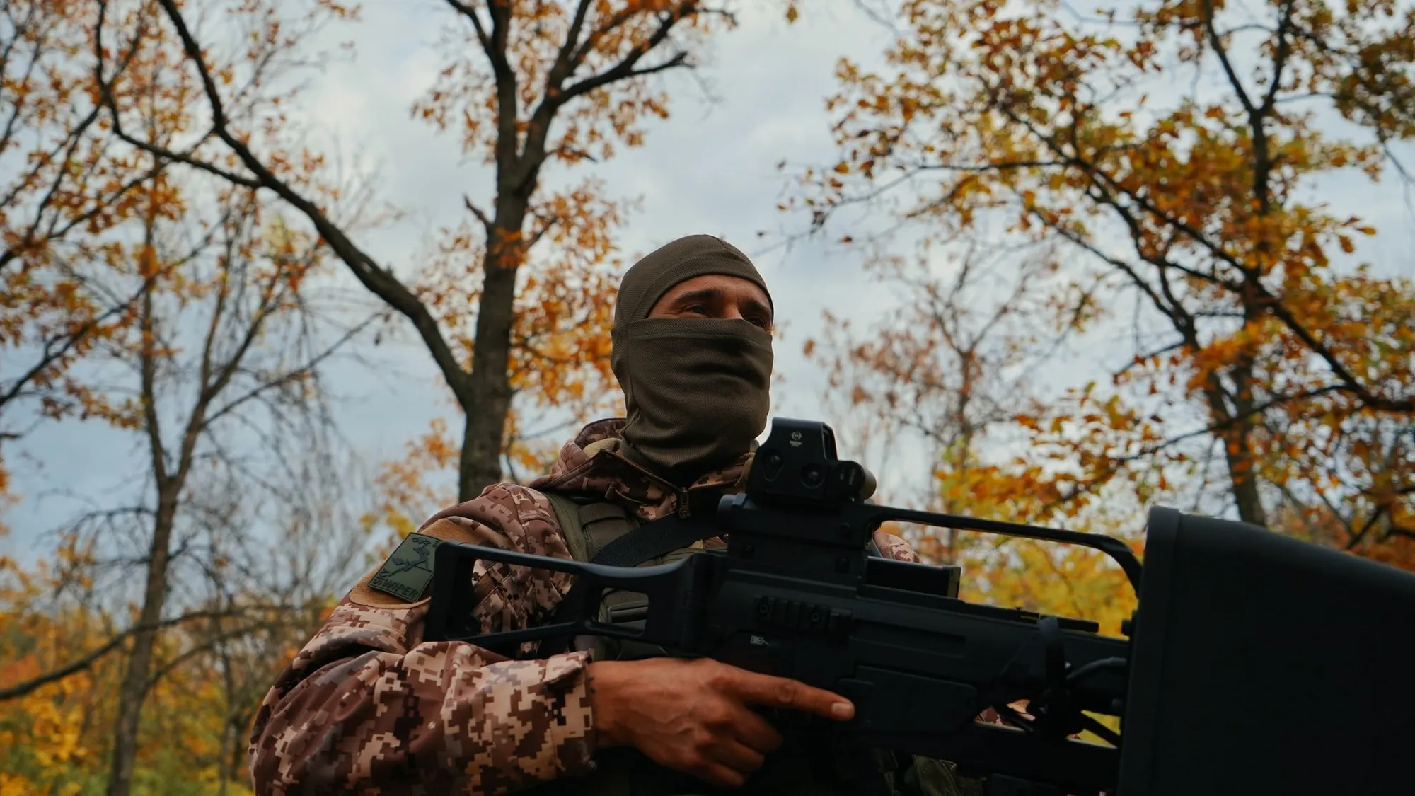 A masked Ukrainian soldier in autumn woodland holding an EDM4S anti-drone jamming gun with a large flat antenna