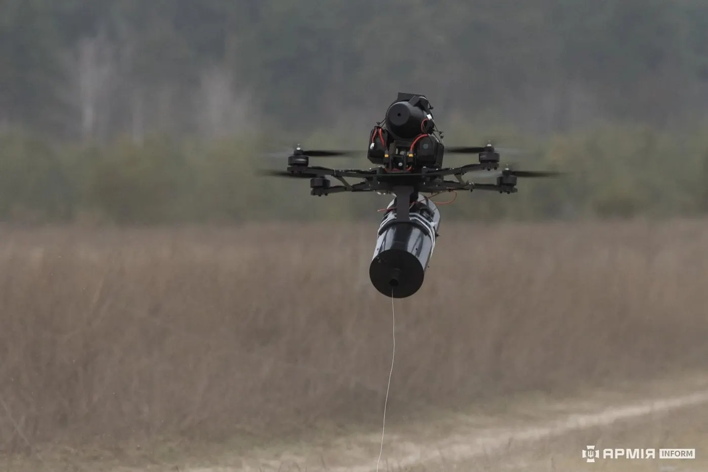 A Ukrainian FPV quadcopter hovering low over dry grass with a cylindrical fibre-optic spool beneath it and a thin cable trailing to the ground