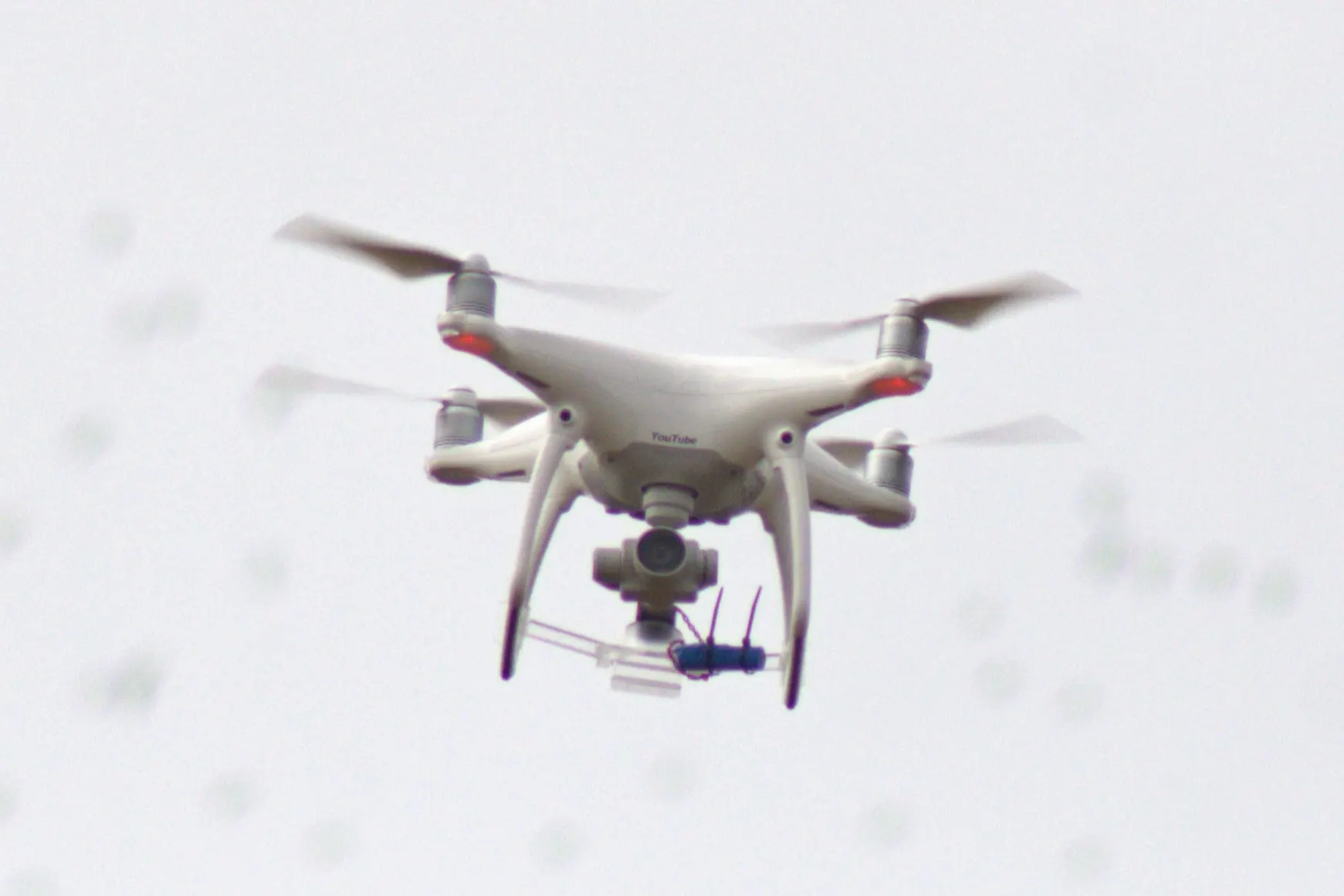 A white DJI Phantom 4 quadcopter with a camera gimbal flying against a pale overcast sky