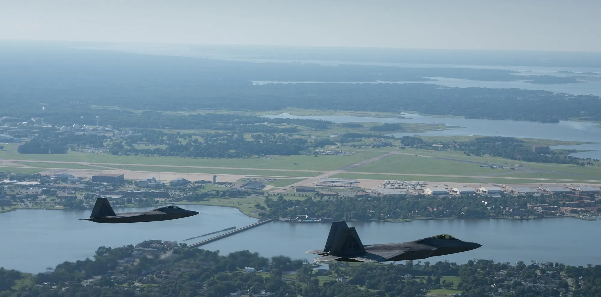 Two U.S. Air Force F-22 Raptors flying low over Joint Base Langley-Eustis in Virginia, with the base and waterways below