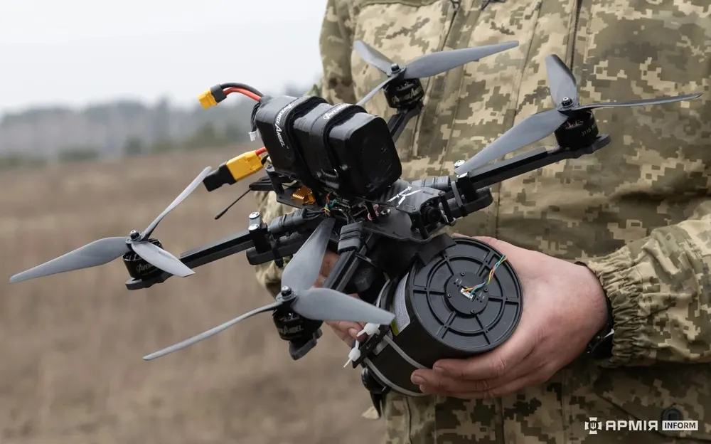 A soldier holds an FPV quadcopter with a black cylindrical fibre-optic cable spool fixed underneath