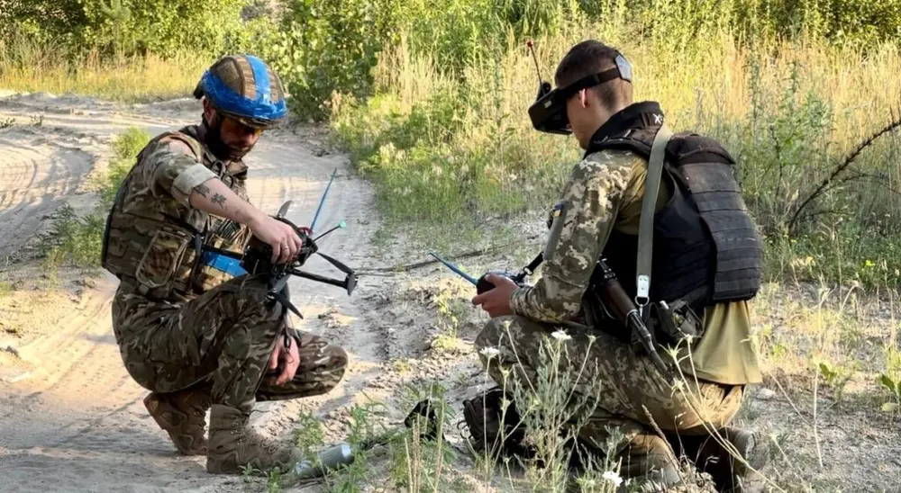 Two Ukrainian soldiers kneel on a sandy track, one wearing FPV goggles, preparing small strike quadcopters