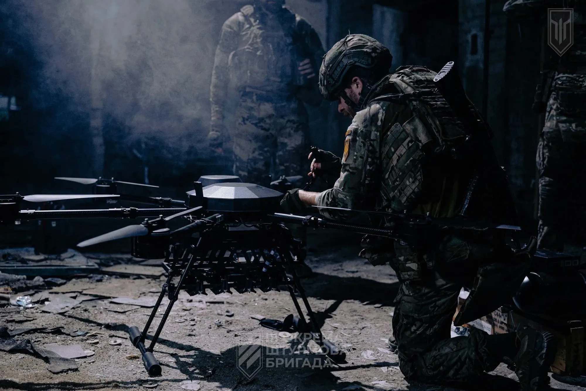 A Ukrainian soldier kneels beside a large six-rotor heavy bomber drone in a dark, smoky ruined building