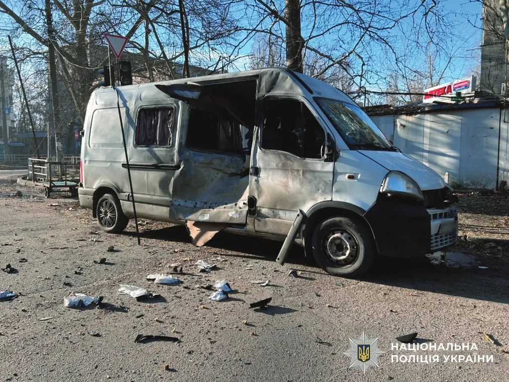 A grey civilian delivery van with its side and windows blown out by a drone strike, parked on a debris-strewn street in Kherson