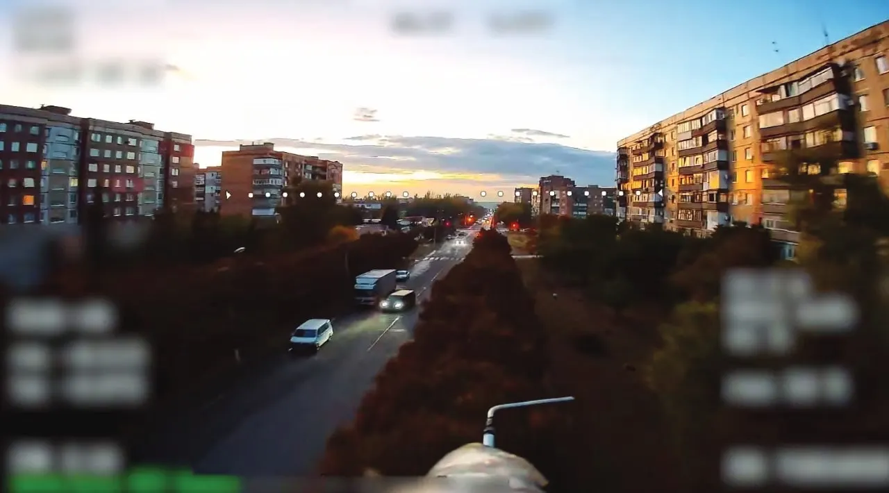 A view from a Russian fibre-optic FPV drone's camera as it flies low between high-rise apartment blocks at dusk, with on-screen data blurred