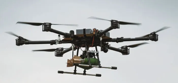A large six-rotor heavy bomber drone with a payload slung underneath, flying against a pale sky