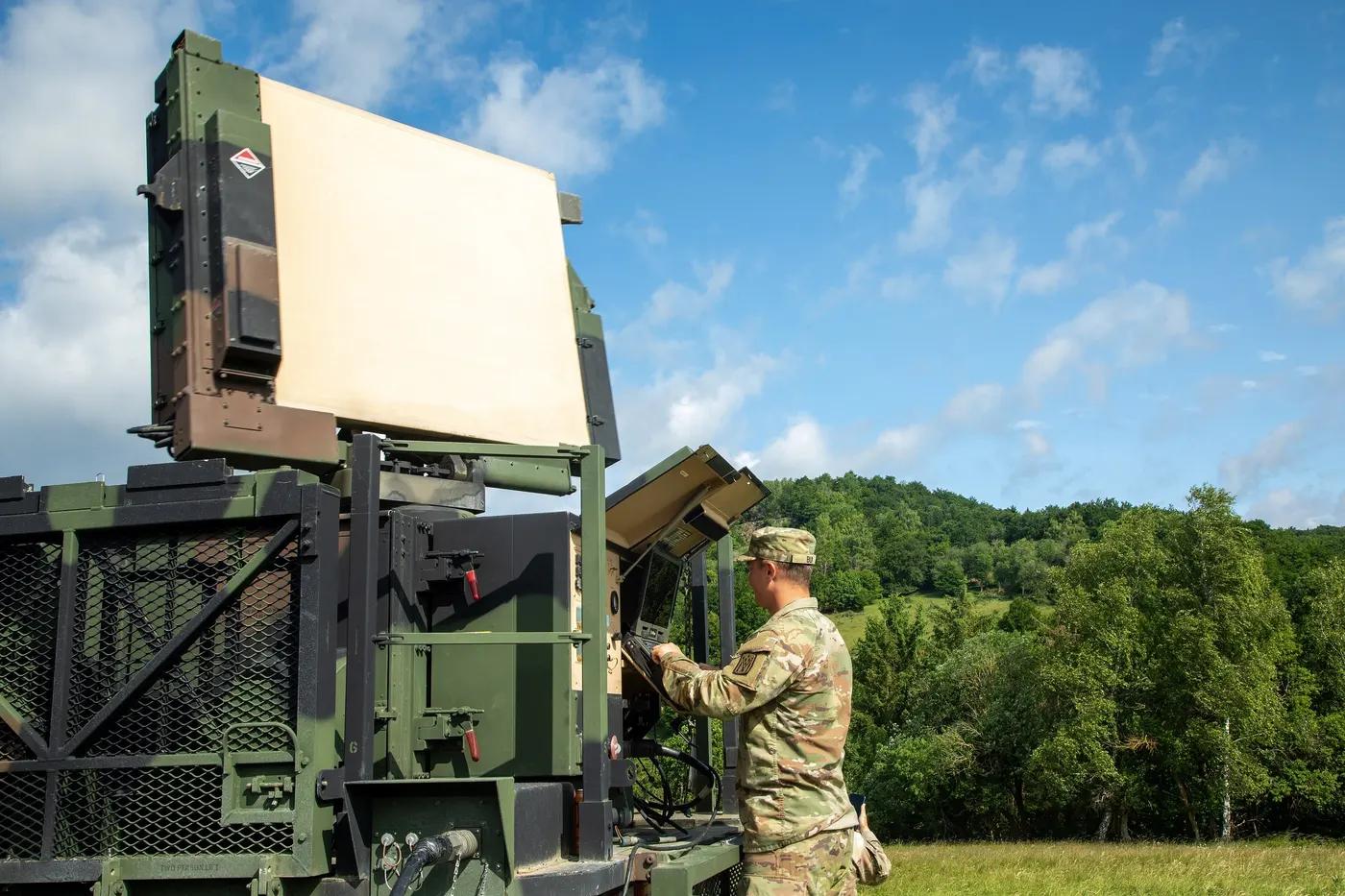 A U.S. soldier operates a trailer-mounted Sentinel radar with a flat rectangular antenna, on a grassy training area under a blue sky