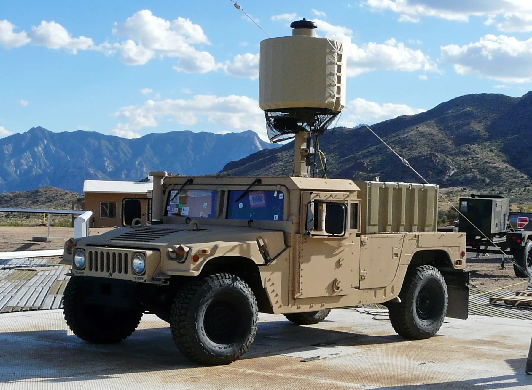 A desert-tan Humvee carrying a cylindrical AN/TPQ-50 radar on a mast, with rugged mountains behind it