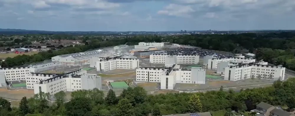 Aerial view of HMP Fosse Way, a modern prison of white multi-storey blocks and exercise yards surrounded by fences, trees and housing